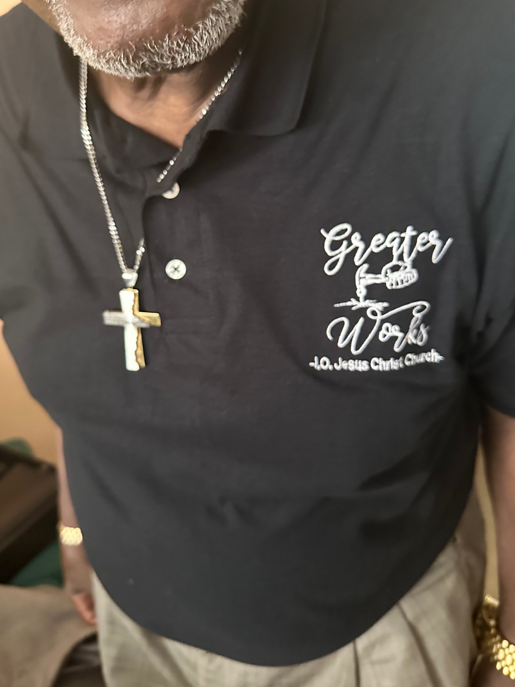 A close-up of a man wearing a black short-sleeve polo shirt with a white printed graphic on the left chest that reads 'Greater Works - La Jolie Catholic Church.' The photo is cropped from the lower face to the waist, showing a silver cross necklace, gold bracelets, and khaki pants in an indoor setting.